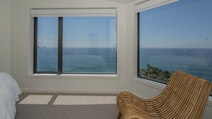 A room with a large window overlooking the ocean, a wicker chair, and a bed with a white comforter.