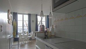 A modern kitchen with white cabinets, a stainless steel countertop, and a dining area with a table and chairs. - Ideally-central cosy & calm studio close to Sainte Catherine - Studio Le Bois Flotté ★★★★ (Honfleur)