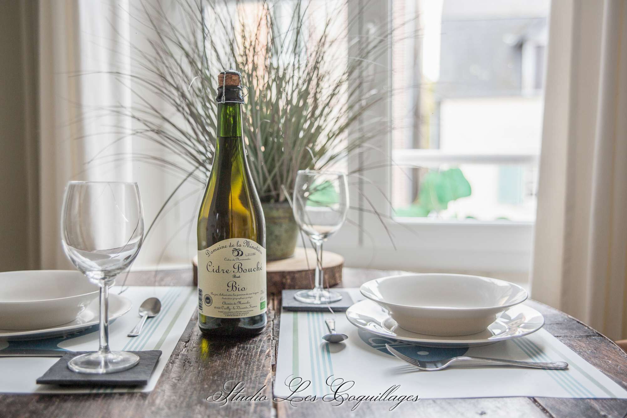Uma garrafa de vinho Sidra Bio Domaine de la Minotiere Bouché, dois copos de vinho e uma mesa de jantar posta com pratos e talheres.