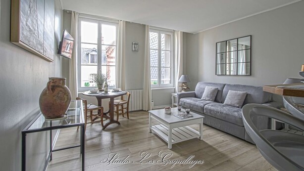 A living room with a grey sofa, a white coffee table, and a dining table with chairs. There is a large window providing natural light and a view of the outside.