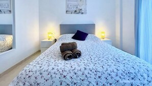 A neatly made bed with a floral bedspread, a rolled-up towel, and a purple pillow. There are two bedside lamps and a framed picture on the wall above the bed. - SHR002 Nuevo y Moderno apartamento en Nerja con Patio (Nerja)