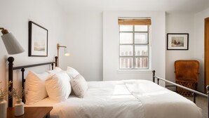 Una cama bien hecha con ropa blanca, una lámpara de noche y un cuadro en la pared.