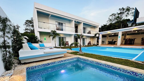 Ein moderner Poolbereich mit einem klaren, blauen Pool, einer weißen Sitzgruppe im Freien mit blauen Kissen und einem zweistöckigen Gebäude mit Balkonen.