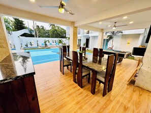 Comedor con mesa de billar, sillas, ventilador de techo y vistas a una piscina y zona de estar exterior.