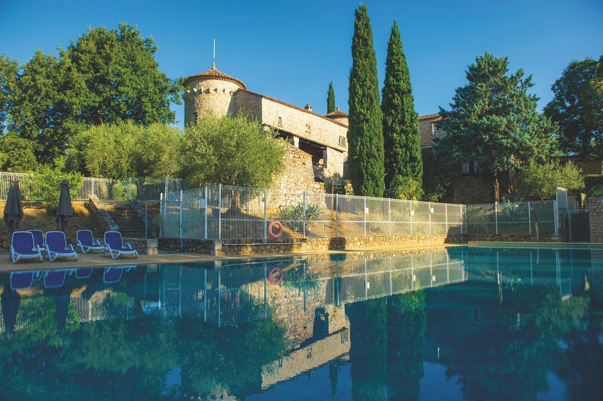 A swimming pool with lounge chairs, a stone building with a tower, and tall trees.