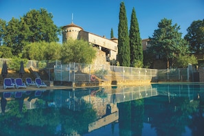 Uma piscina com espreguiçadeiras, um edifício de pedra com torre e árvores altas.