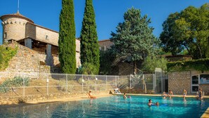 Ein Schwimmbecken mit Personen, ein Steinhaus mit einem Turm und hohe Bäume.