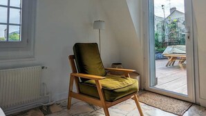 Huone, jossa on korkea katto, suuri ikkuna, pehmustettu tuoli, lattiavalaisin ja näkymä ulkoterasille.