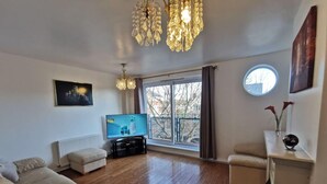 Un salon comprenant une télévision, un canapé, une table basse, un lustre et une grande fenêtre donnant sur la forêt.
