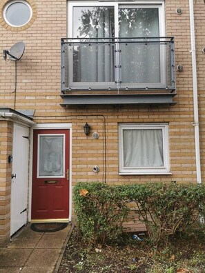 A two-story building with a red door, a small balcony with a metal railing, and a window with a curtain.
