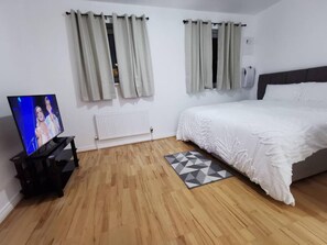 A bedroom with a bed, a TV on a stand, two windows with curtains, a radiator, and a geometric rug.