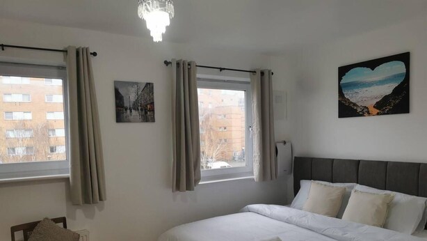 Une chambre à coucher avec un lit, deux fenêtres avec des rideaux, une chaise, un radiateur et un tableau accroché au mur.