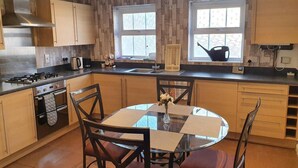 A modern kitchen with wooden cabinets, a gas stove, a sink, and a dining area with a glass table and chairs. - Mannys Apartment - Flagship Lodge (Sittingbourne)