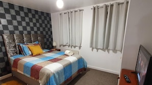 A bedroom with a bed, a headboard, curtains, a radiator, a remote control, and a television. - Mannys Apartment - Flagship Lodge (Sittingbourne)
