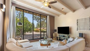 A living room with a large window, a ceiling fan, a sofa, a glass coffee table, and a flat-screen TV. - Sedona Modern Mid-Century Walk to Hiking Trails, Natural Light, Dinner In The Yard! (Sedona)