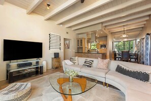 Una sala moderna con un televisor de pantalla plana grande, una mesa de centro de vidrio, un sofá color beige y una cocina al fondo.