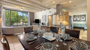 A dining area with a large table set for six, surrounded by chairs, with a kitchen in the background. - Sedona Modern Mid-Century Walk to Hiking Trails, Natural Light, Dinner In The Yard! (Sedona)