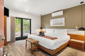 Un dormitorio moderno con una cama grande, mesitas de noche, un banco de madera, un equipo de aire acondicionado y una puerta corrediza de vidrio que da a un espacio al aire libre.