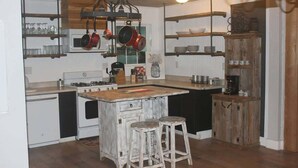 Une cuisine dotée d’un îlot central, de tabourets en bois, de casseroles suspendues et d’étagères où sont rangées assiettes et tasses.