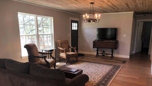 Un salon spacieux avec un grand canapé, deux fauteuils, une table basse en bois et un téléviseur posé sur un meuble. Un lustre est suspendu au plafond, et une grande fenêtre laisse entrer la lumière naturelle.