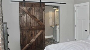 Une chambre à coucher avec un lit, une porte coulissante en bois, une salle de bain avec un lavabo et un miroir, et une porte blanche.