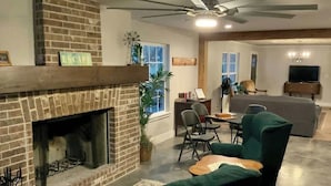 Un salon avec une cheminée en briques, des fauteuils verts, une table à manger et un ventilateur de plafond.