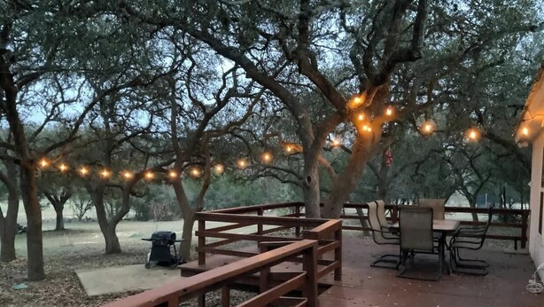 Une terrasse en bois avec une table de pique-nique et des chaises, entourée d’arbres et illuminée de guirlandes lumineuses.