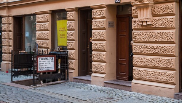 Fachada de un edificio con un letrero que indica Kredis Pub, una puerta con una placa con el número 10 y un ventanal con un letrero amarillo.