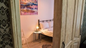 Un dormitorio con cama, una silla, una mesita de noche con lámpara y un cuadro en la pared.