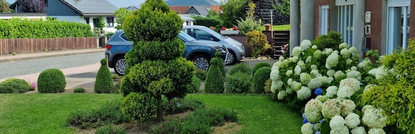 Una entrada de casa con un árbol podado, un coche aparcado y un jardín bien cuidado con hortensias.