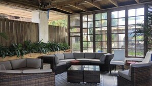 A covered outdoor seating area with wicker furniture, large windows, and potted plants.