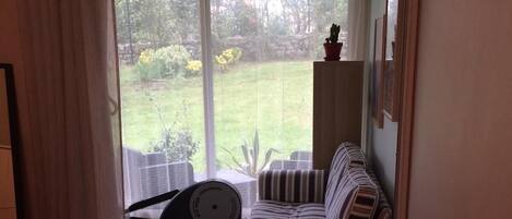 A bedroom with a bed, a striped chair, a small table with a lamp, and a view of a garden through a window.
