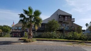 Un edificio de dos pisos con un balcón en el nivel superior, rodeado de vegetación y una bandera.
