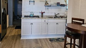 Una cocina con armarios blancos, estufa a gas, lavamanos y ventilador de techo. Hay una mesa de comedor con sillas y una alfombra en el suelo.