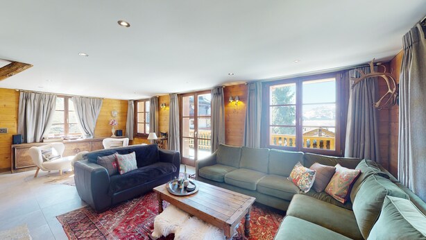 A spacious living room with a dark blue sofa, a light green sofa, and a wooden coffee table. There are large windows with curtains, a wooden wall, and a view of a building outside. - Charming Chalet - Walk to Piste & Villars Centre! Style & Comfort 5 Bed/4 Bath! (Ollon)