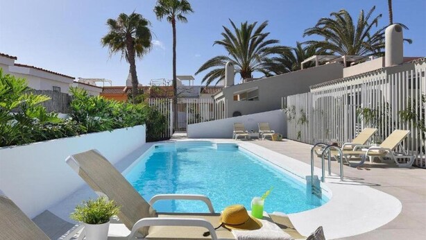 A swimming pool with lounge chairs, palm trees, and a white fence. - Velsuites (San Bartolome de Tirajana)