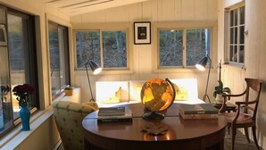 A cozy room with a wooden ceiling, a round table with books and a globe, a chair, a vase with flowers, and a window with a view of trees. - Historic Pound Ridge Cottage, 2 Beds, Fully Furnished (Pound Ridge)
