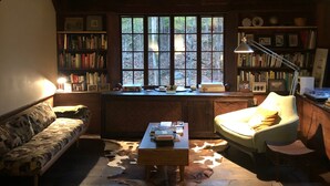 A cozy room with a wooden ceiling, a large window with a view of trees, a bookshelf, a sofa, a chair, and a table with books. - Historic Pound Ridge Cottage, 2 Beds, Fully Furnished (Pound Ridge)