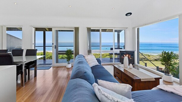 A modern living room with a large sofa, a dining table, and a fireplace. There are large windows offering a view of the ocean. - 5 Whitecrest Resort - Pool & Amazing Ocean Views (Wongarra)