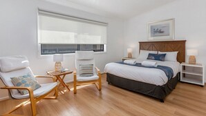 A bedroom with a bed, two chairs, a nightstand, a window with blinds, and a framed picture on the wall. - 5 Whitecrest Resort - Pool & Amazing Ocean Views (Wongarra)