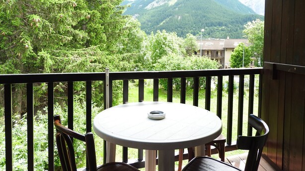 Ein Balkon mit rundem Tisch und Stühlen, der auf eine gebirgige Landschaft mit Bäumen und Gebäuden blickt.