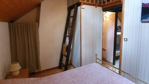 Ein Schlafzimmer mit schrägen Wänden, einem Bett mit floralem Überwurf, einer Leiter, einem Fenster mit Vorhängen und einer Tür zu einem anderen Raum.