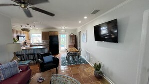 Ein Wohnzimmer mit einer Couch, einem Couchtisch und einem an der Wand montierten Fernseher. Es gibt eine Essecke mit Tisch und Stühlen, und im Hintergrund befindet sich eine Küche.