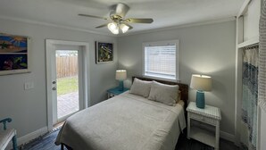 Ein Schlafzimmer mit einem Bett, zwei Nachttischen mit Lampen, einem Deckenventilator, einer zur Außenwelt führenden Tür und einem Fenster mit Jalousien.