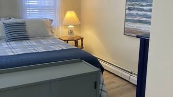 Une chambre à coucher comprenant un lit, une table de chevet, une lampe, un tableau représentant l’océan et une fenêtre avec des stores.