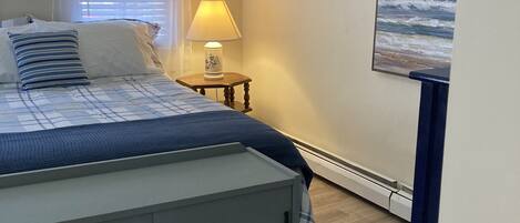 A bedroom with a bed, a nightstand, a lamp, a painting of the ocean, and a window with blinds.