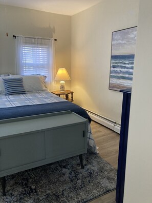 A bedroom with a bed, a nightstand, a lamp, a painting of the ocean, and a window with blinds.