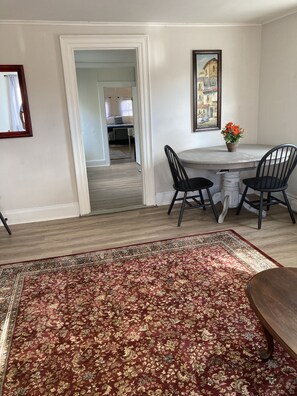 A dining area with a marble table, two black chairs, a red and gold patterned rug, and a framed painting on the wall.