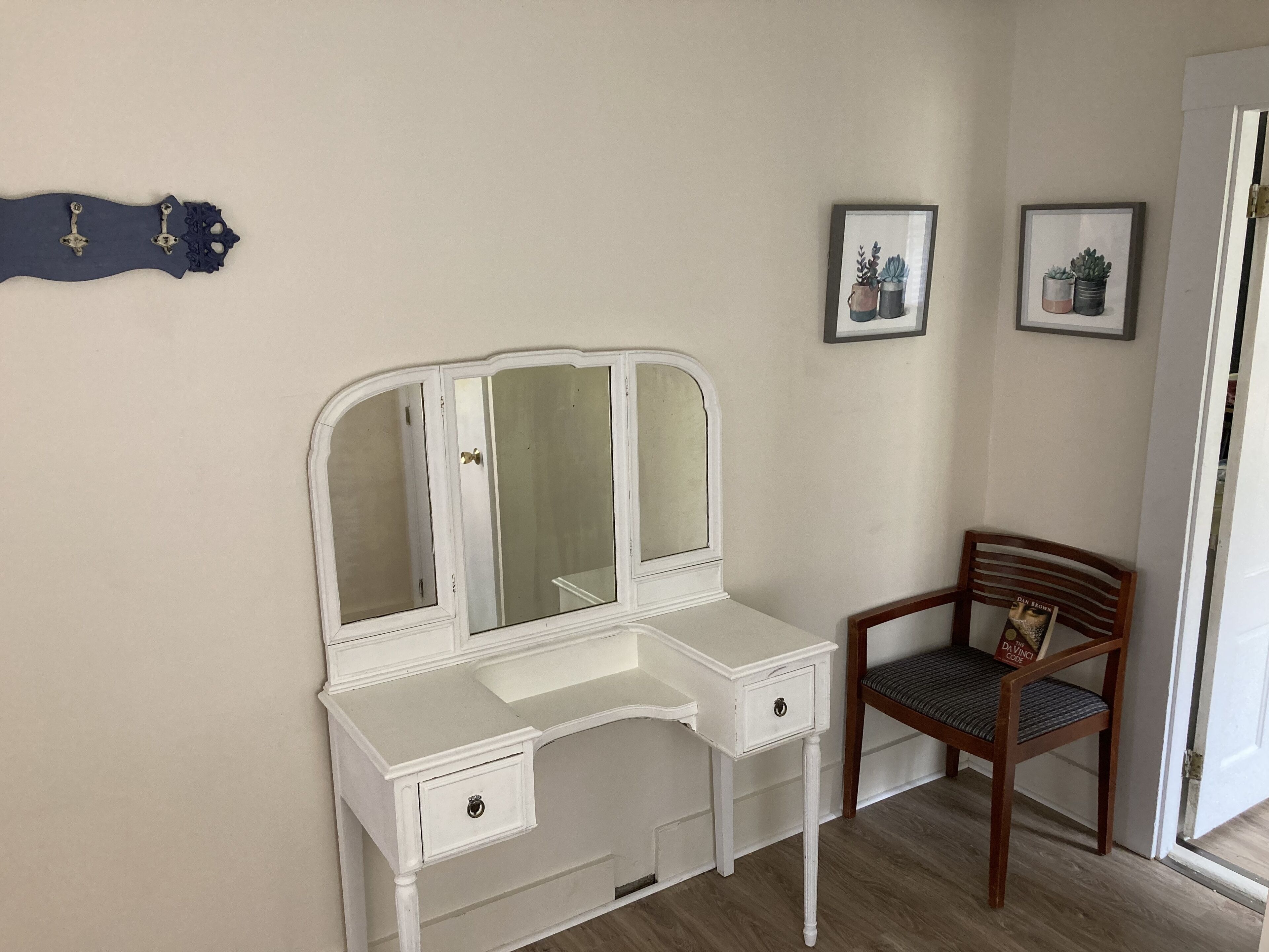 Een witte kaptafel met spiegel, een houten stoel en twee afbeeldingen in lijsten aan de muur.