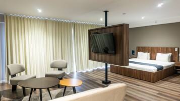 Habitación de hotel con cama, televisión montada en un panel de madera, zona de estar con una silla y una mesita, y una ventana grande con cortinas.